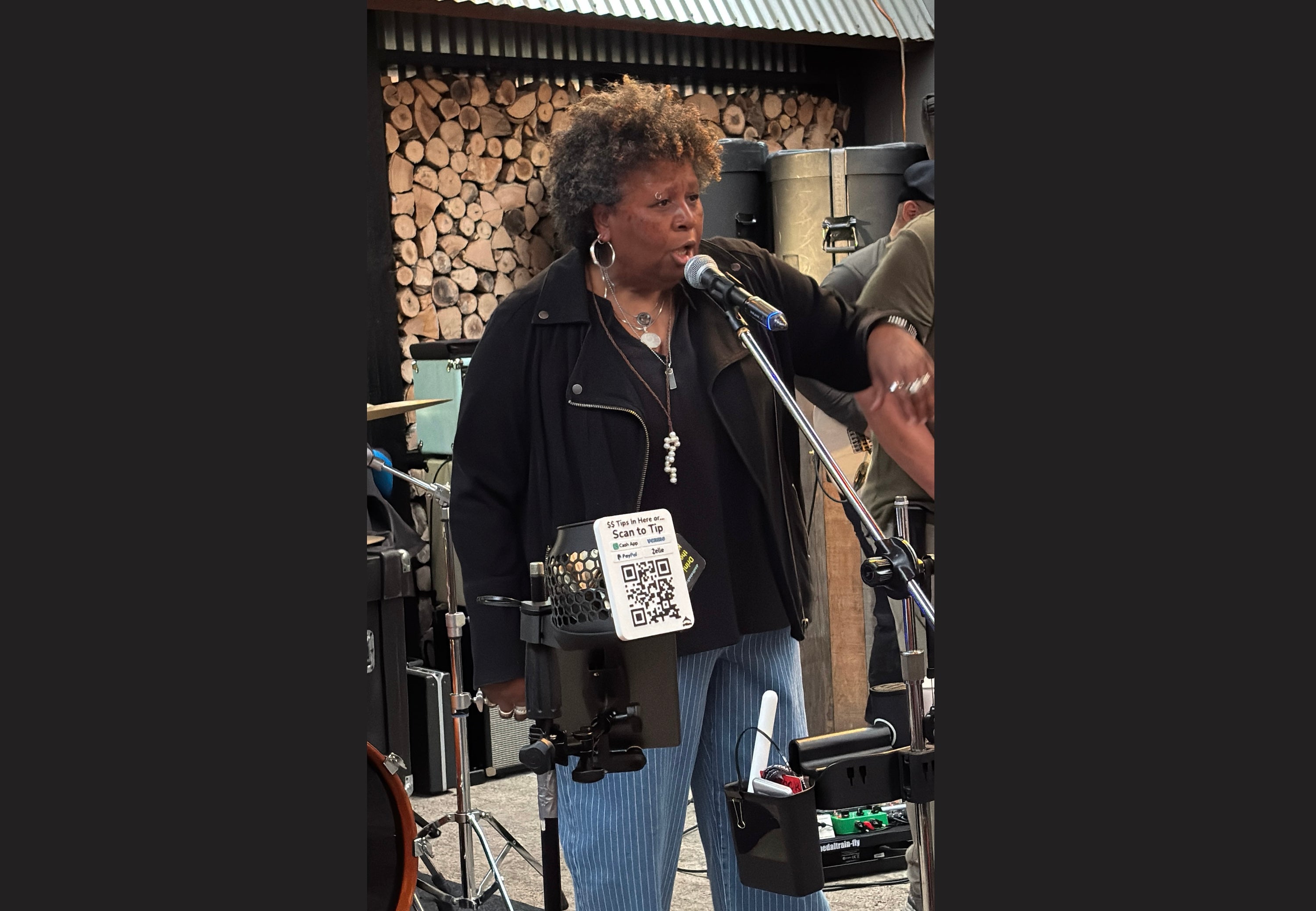 Performer using an Artfare Tip Jar Holder during a live outdoor music performance, showing a QR payment sign for Venmo, Cash App, PayPal, and Zelle mounted to a microphone stand