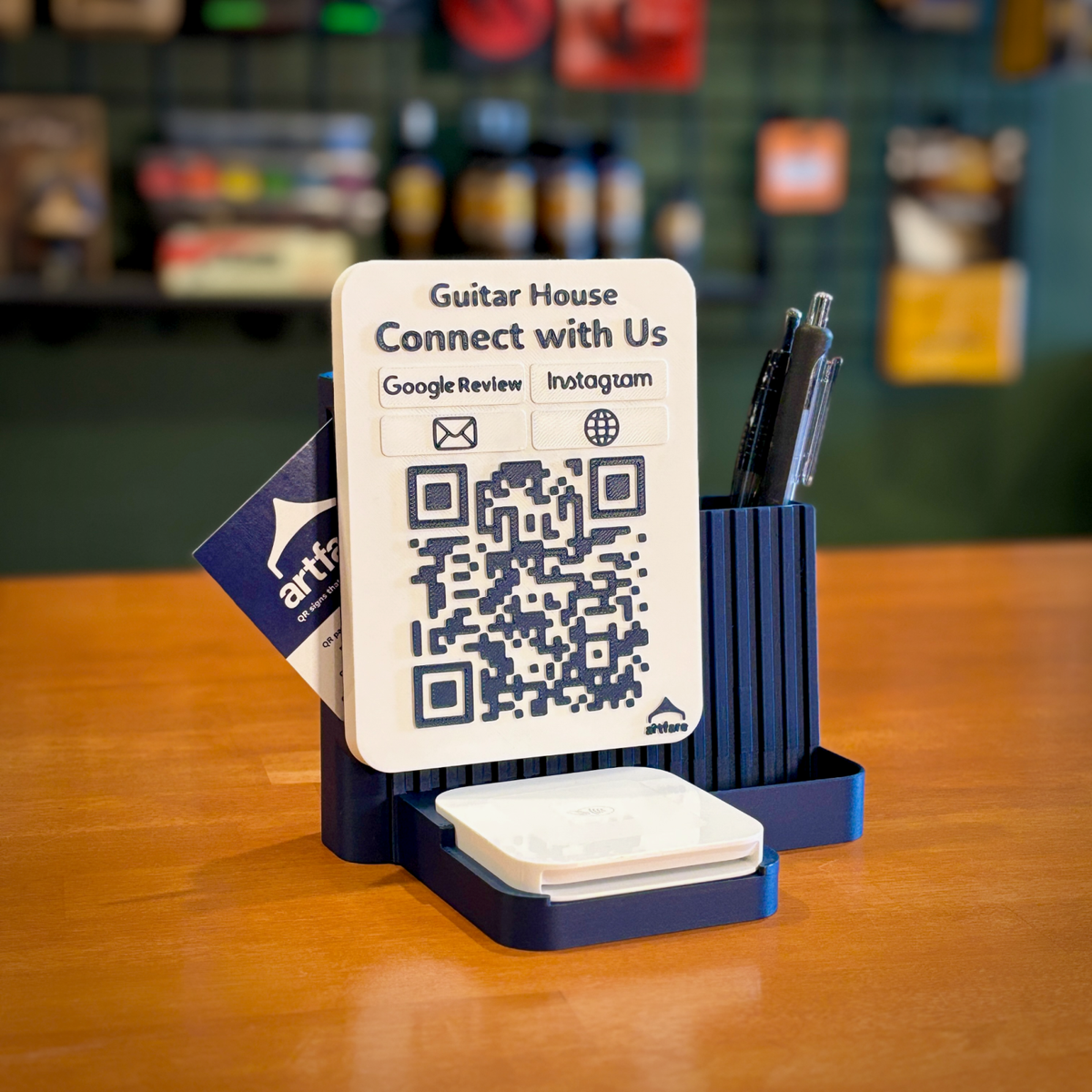 Navy Shopkeeper Square Reader dock with Guitar House QR sign, Square Reader in front, pens and business cards on a shop counter